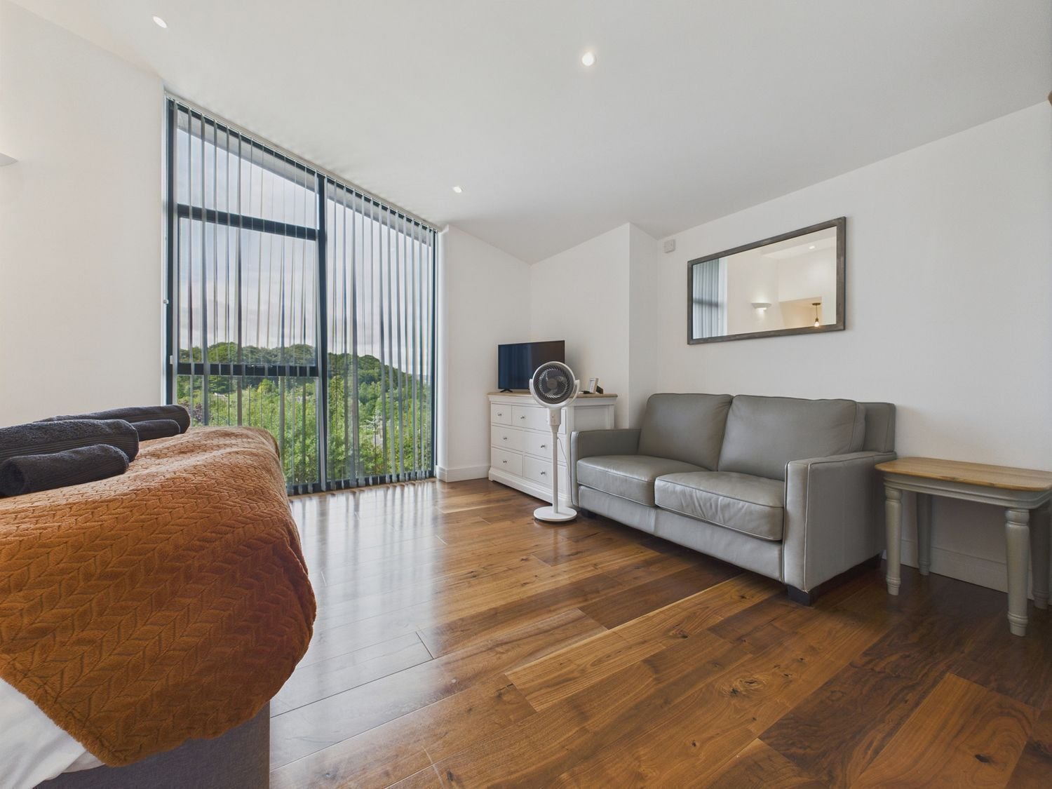 Quarto moderno com cama, sofá cinza, mesa pequena, cômoda e ventilador.