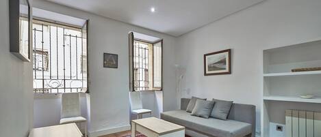 A living room with a sofa, a small table, a chair, and a window with bars.