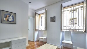 Ein Zimmer mit weißer Wand, einem gerahmten abstrakten Gemälde, einer weißen Bank, einem kleinen Tisch und einem Fenster mit Metallgitter.