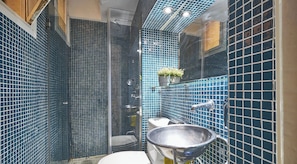 Ein Badezimmer mit blauen Mosaikfliesen, einer Glasdusche, einer Toilette, einem Waschbecken und einem Metallbecken.