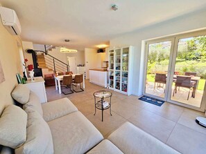 A modern living room with a sofa, a dining area with chairs, a kitchen with cabinets, and a sliding glass door leading to an outdoor patio.