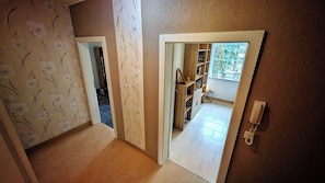 Un couloir avec un papier peint à motifs, une porte en bois et une vue sur une pièce dotée d’une fenêtre et d’étagères.