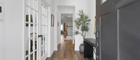 A hallway with white walls, wooden flooring, and a dark door. There is a potted plant and a framed picture on the wall.