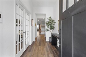 Un couloir aux murs blancs, doté d’un plancher en bois et d’une porte sombre. On y trouve une plante en pot et un tableau encadré accroché au mur.