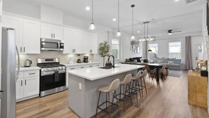 A modern kitchen with white cabinets, stainless steel appliances, a central island with bar stools, and pendant lighting. - NRG Access | Sleeps 10 | Near The Heights (Houston)