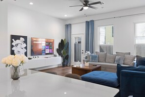 Ein modernes Wohnzimmer mit einem weißen Sofa, blauen Sesseln, einem Couchtisch und einem Fernseher, der Streamingdienste anzeigt.