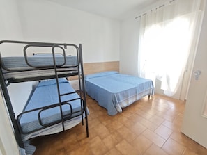 Ein Schlafsaalzimmer mit Etagenbetten, einem Holz-Kopfstück, einem Fenster mit durchsichtigen Vorhängen und einem gefliesten Boden.