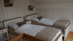 Quarto com duas camas de madeira, roupa de cama branca, mesa de cabeceira com abajur e um arranjo floral fixado na parede.