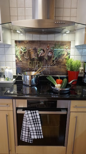 Une cuisine dotée d’une hotte en acier inoxydable, d’un pare-feu en bois sur lequel est inscrit le mot « GUTEN », ainsi que de divers ustensiles de cuisine et de produits frais disposés sur le plan de travail.