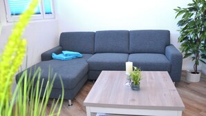 Un sofá seccional gris, una mesa de centro de madera, una planta pequeña en maceta y una ventana con persianas.