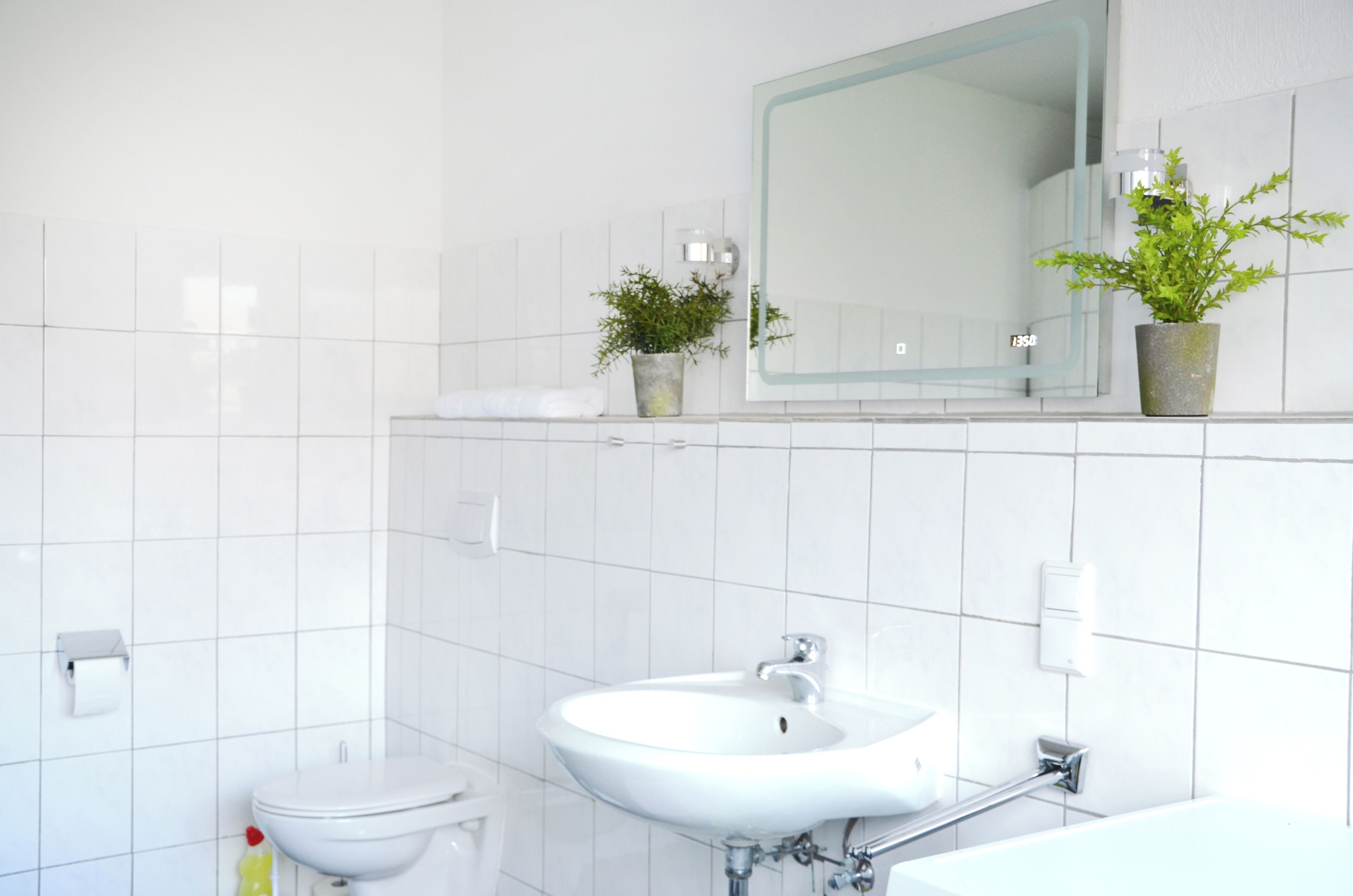 A bathroom with white tiles, a sink, a toilet, and two potted plants.