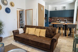 Ein Wohnzimmer mit braunem Sofa, einem Esstisch mit Stühlen, einer Küche mit dunklen Schränken und einer Topfpflanze.