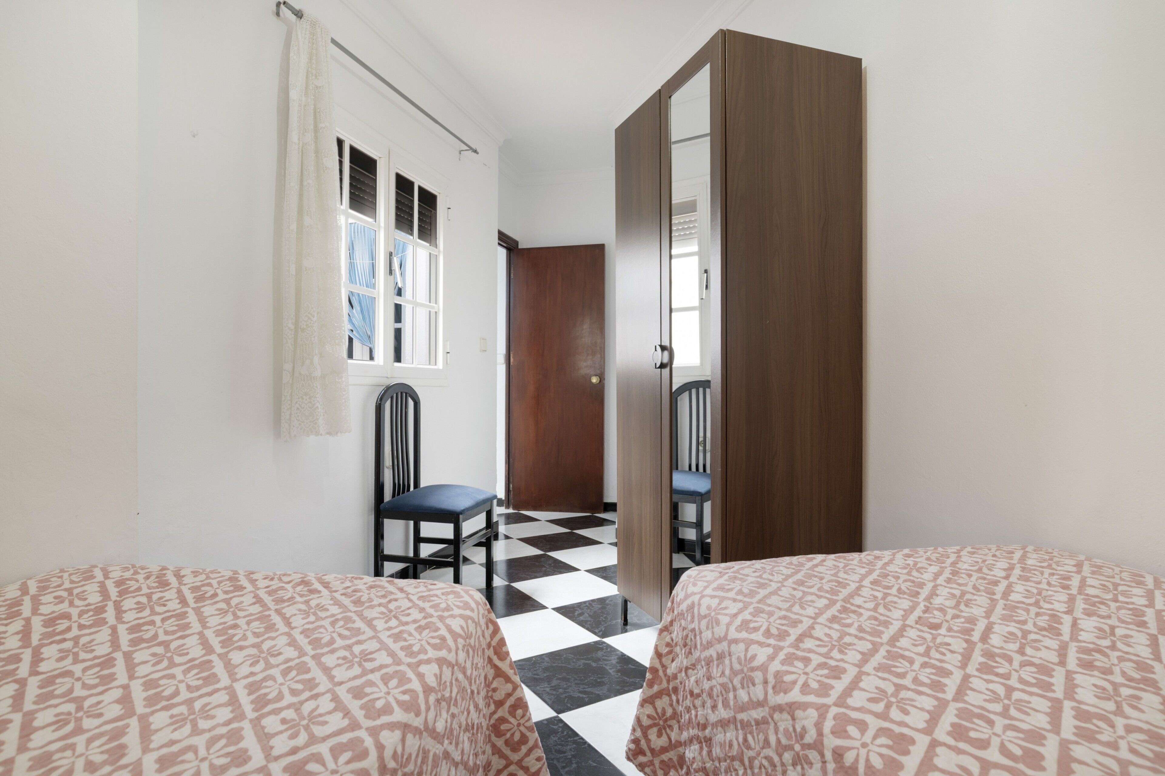 A room with two beds, a chair, a wooden wardrobe, a window with a curtain, and a checkered floor.