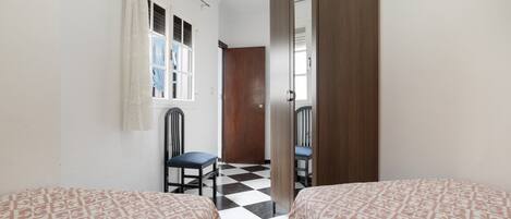 A room with two beds, a chair, a wooden wardrobe, a window with a curtain, and a checkered floor.