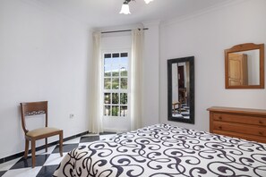 Ein Schlafzimmer mit Schachbrettmuster-Boden, einem Bett mit gemustertem Überwurf, einem Holzstuhl, einer Kommode, einem Spiegel und einem Fenster mit Vorhängen.