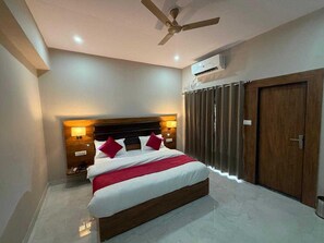 Una habitación de hotel con cama, ventilador de techo, aire acondicionado, una puerta de madera y una mesita de noche con lámpara.
