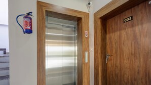 Una puerta de madera con una placa numérica, un extintor de incendios colgado en la pared y una puerta de ascensor de vidrio.