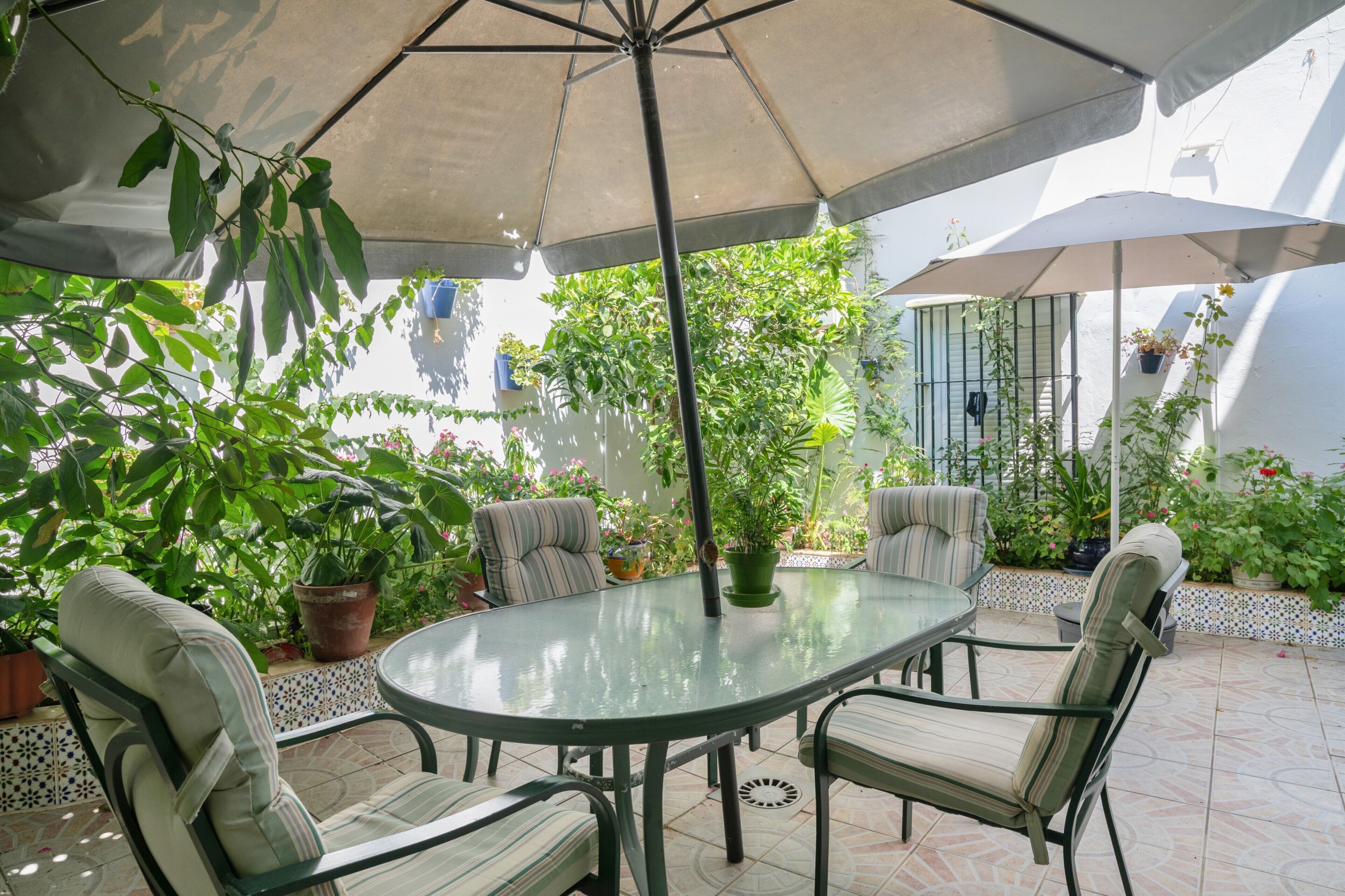 Une terrasse extérieure avec une table en verre, quatre chaises et deux parasols pour créer de l’ombre.