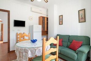Una sala de estar con sofá verde, sillas de madera, una mesa de vidrio con un tazón azul, refrigerador blanco, televisor de pantalla plana y un reloj en la pared.