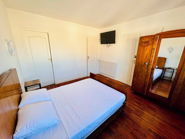Une chambre avec un lit en bois, une petite table de chevet, un miroir, un téléviseur fixé au mur et un radiateur.