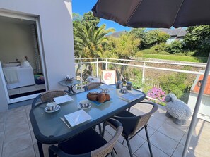 Ein Balkon mit Tisch für zwei Personen, einem Korbstuhl, einer Statue und Blick auf Grünflächen und ein Haus.