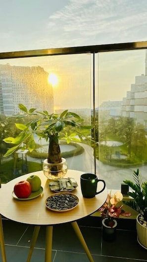Un balcón con una barandilla de vidrio, una planta en maceta, una mesa con frutas, una taza y un adorno.