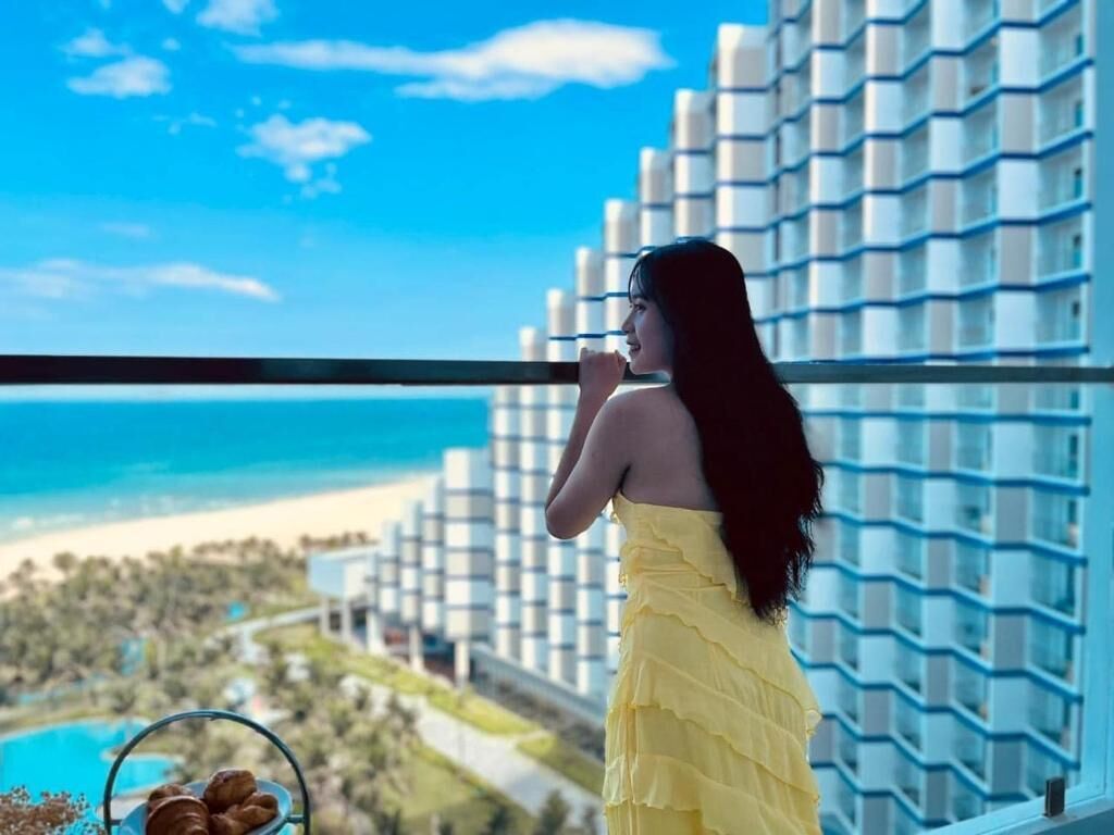 Uma pessoa de vestido amarelo está num terraço com vista para uma praia e um edifício moderno.
