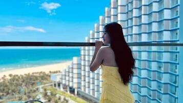 Uma pessoa de vestido amarelo está num terraço com vista para uma praia e um edifício moderno.