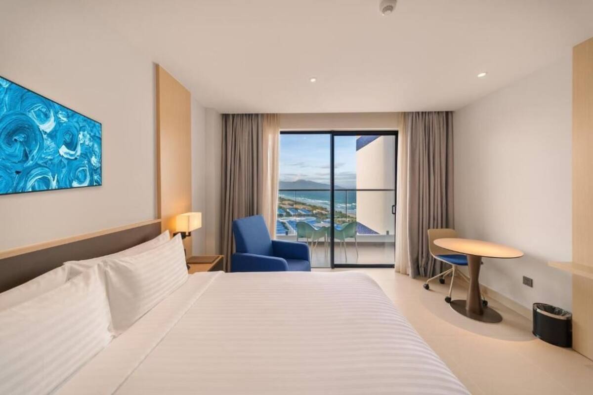 Habitación de hotel con cama, una silla, una mesa y vista al océano a través de un ventanal grande.
