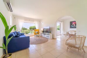 Ein Wohnzimmer mit einem blauen Sofa, einem gelben Sessel, einem Fernseher und einem Essbereich mit Stühlen.