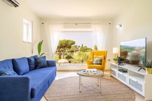 Ein Wohnzimmer mit blauem Sofa, geläufigem Sessel, Fernseher und Blick ins Grüne.