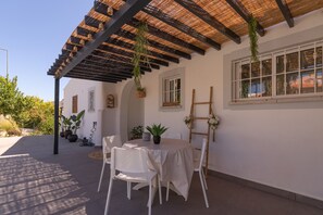 Eine Terrasse im Freien mit Tisch und Stühlen, eine Pergola mit hängenden Pflanzen und ein Fenster mit Blick auf die Umgebung.