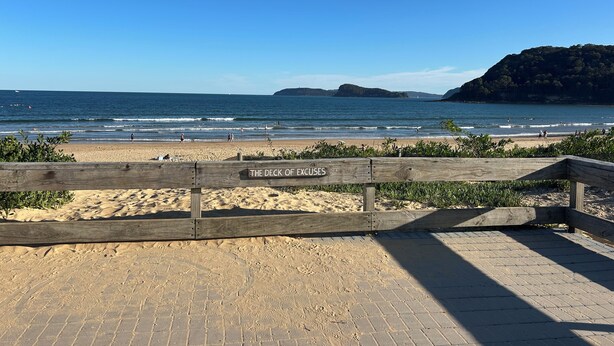 Uma praia com uma cerca de madeira com a inscrição 'The Deck of Excuses', uma faixa de areia e o oceano.