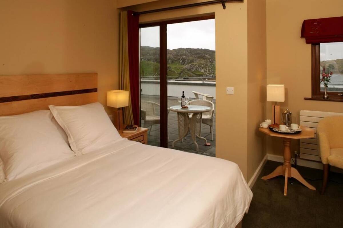 Quarto de hotel com uma cama, uma mesinha com luminária, uma cadeira e vista para o exterior.