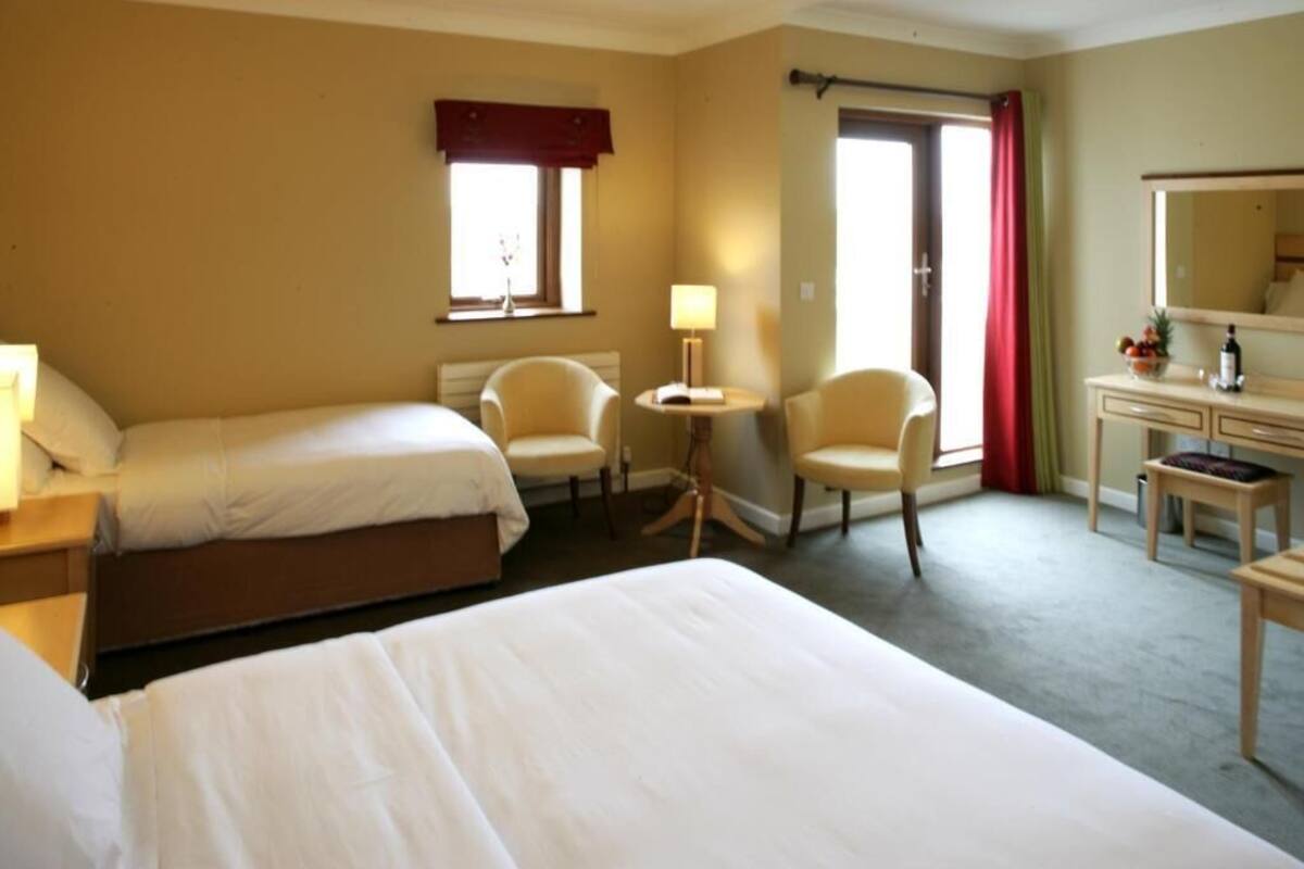 Quarto de hotel com duas camas, uma mesa, uma cadeira, um abajur e uma janela com cortinas.