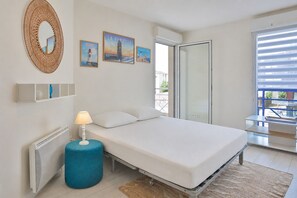 Ein Schlafzimmer mit einem Bett, einem runden blauen Hocker, einem Heizkörper, einem an der Wand befestigten Spiegel und einer Tür, die auf einen Balkon führt.