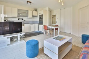 Eine moderne Küche mit weißen Schränken, eine Essecke mit Tisch und Stühlen, ein Fernseher und ein blauer Hocker.