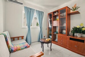 Ein Wohnzimmer mit einer Couch, einem kleinen runden Tisch, einer Holzkommode und einem Fenster mit Vorhängen.
