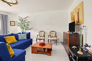 Ein Wohnzimmer mit einem blauen Sofa, zwei Sesseln, einem Couchtisch, einem Fernseher, einem Schrank und einer Pflanze.