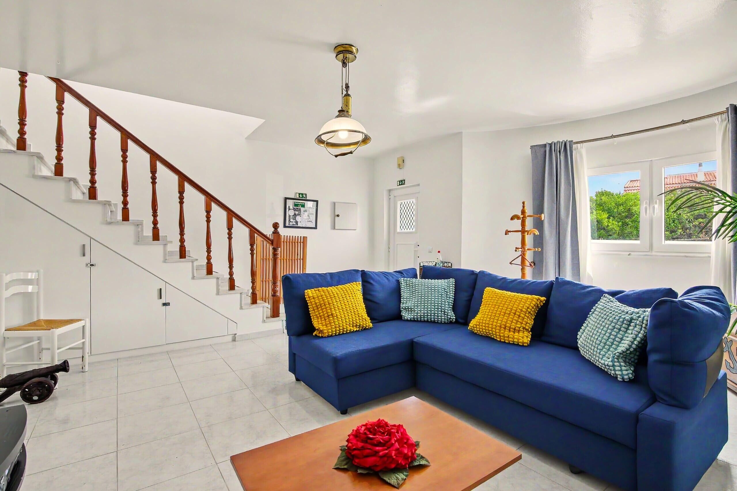 Una sala de estar con un sofá azul, una escalera de madera, una lámpara en el techo y una ventana con cortinas.