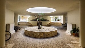 Ein kreisrunder Innenhof mit Steinplatten und einem zentralen Baum, umgeben von Stufen, die zu einer erhöhten Ebene führen.