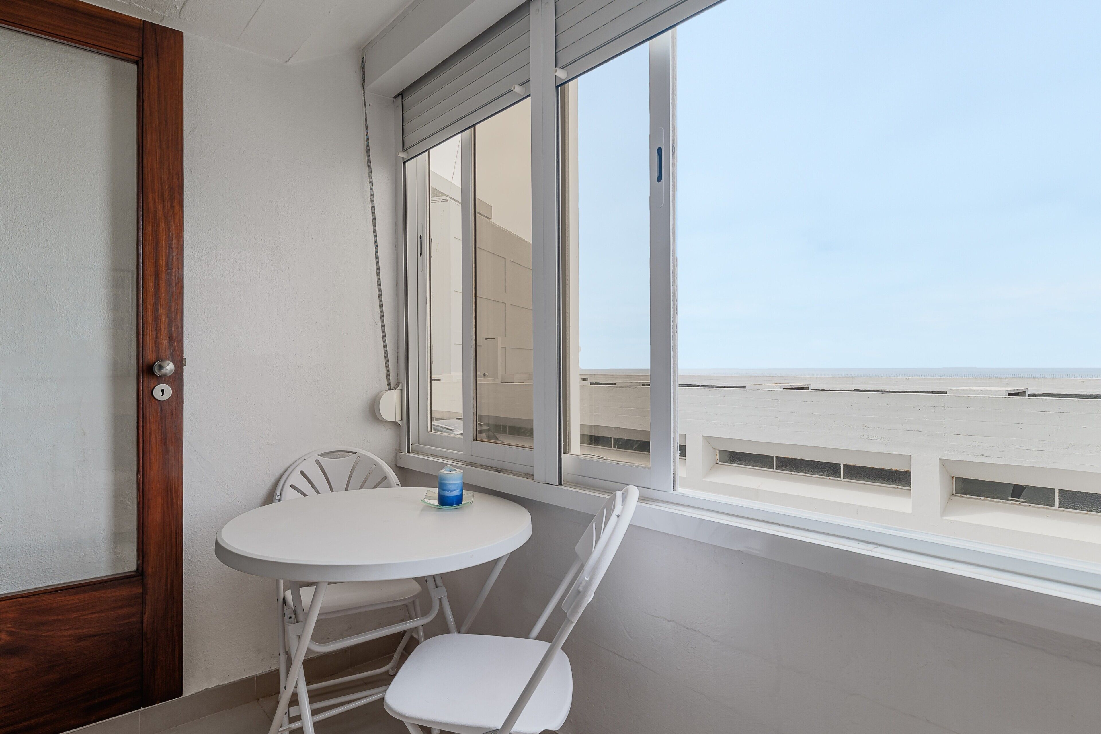Ein Balkon mit weißem Tisch und Stühlen, ein Fenster mit Blick auf den Strand und eine Tür mit Holzrahmen.
