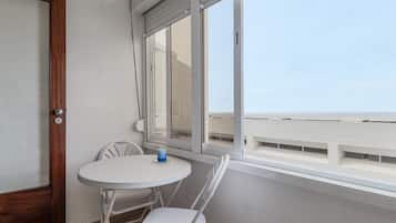 Uma varanda com uma mesa e cadeiras brancas, uma janela com vista para a praia e uma porta com moldura de madeira.