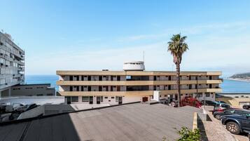 Um hotel com vários andares, área de estacionamento, uma palmeira e vista para o mar.