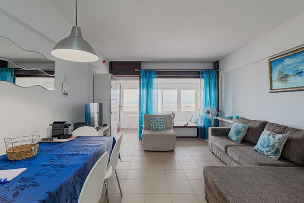 A modern living room with a dining area, a sofa, a television, and a view of the sea through large windows.