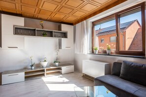 A modern living room with built-in shelves, a glass coffee table, and a view of a red-roofed building.