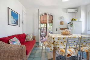 Un coin repas avec une table recouverte d’une nappe à motifs de tournesols, un canapé rouge et une chaise en osier.