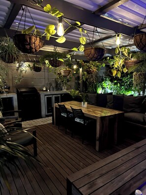 Eine Holzterrasse mit Tisch und Stühlen, umgeben von hängenden Pflanzen und einem Grill.