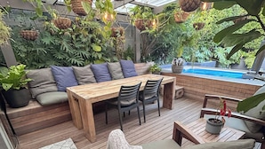 Eine überdachte Terrasse im Freien mit einem Holztisch, Stühlen, einer Bank mit Kissen, hängenden Pflanzgefäßen und einem Pool.