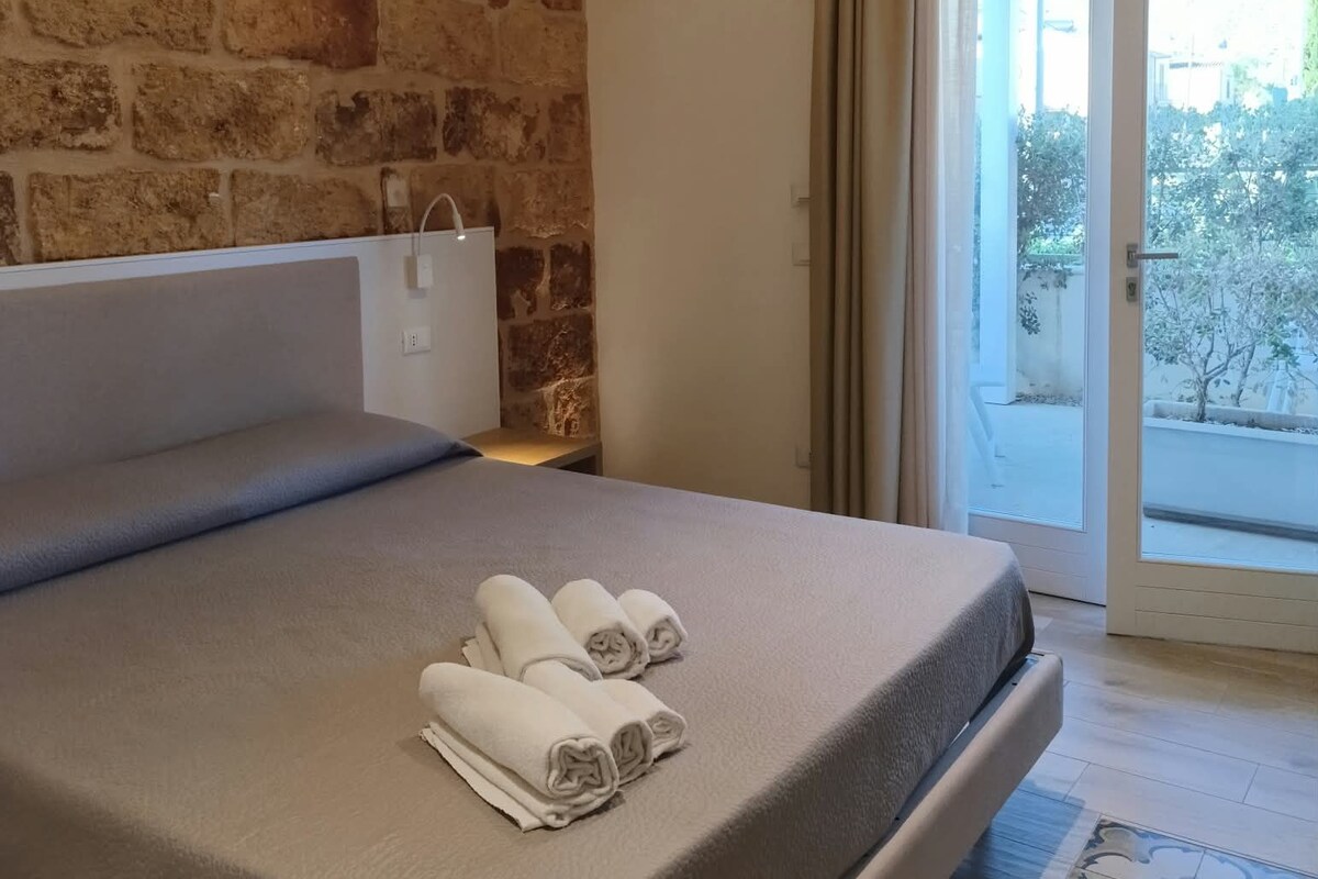 A bedroom with a stone wall, a bed with a grey mattress, and a view of the outdoors through a sliding door.